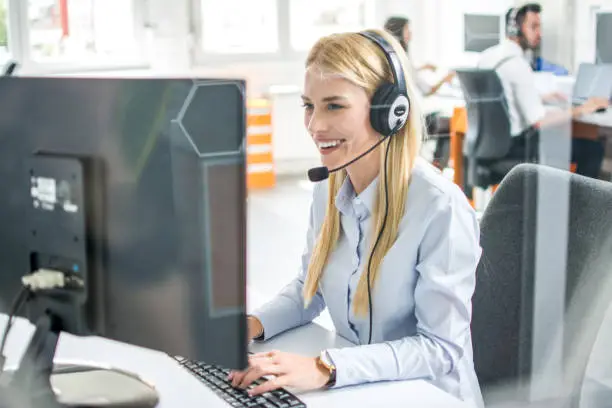 Photo of Friendly smiling customer support agent woman talking with client using headset and typing on computer in call center.