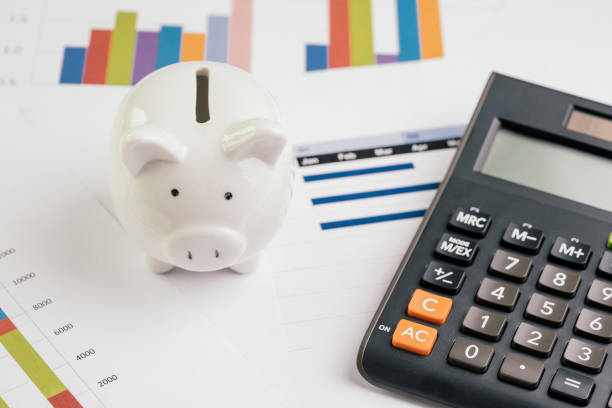 Savings and investment concept, white piggy bank with black calculator on pile or economic analysis report chart and graph on office table stock photo