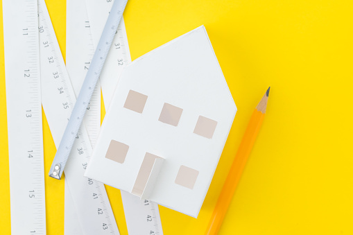 White paper house, yellow pencil and white measuring tapes with centimetre and inches on vivid yellow background, architecture, house building and renovation.