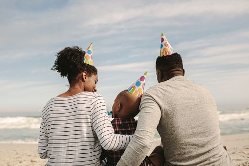 Rear view of african family with party hats blowing whistle and having fun on the beach. Man and woman celebrating son's birthday.
