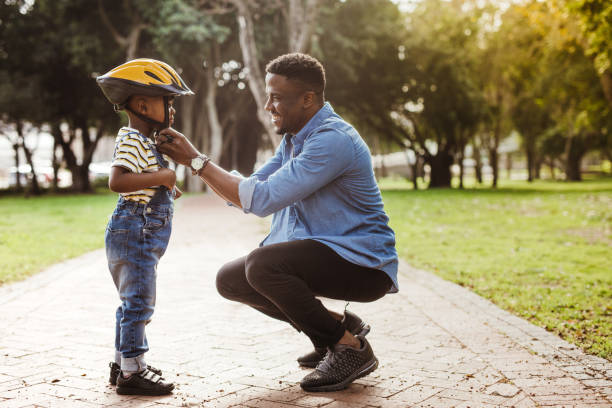 Father puts his son a helmet for riding bike African man putting helmet on cute boy at the park. Father puts his son a protective helmet for riding bike. cycling helmet photos stock pictures, royalty-free photos & images