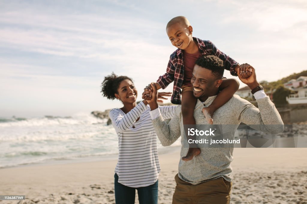 Parents carrying son on shoulders on beach vacation Parents carrying son on shoulders on beach vacation. African family of mother and father carrying son on his shoulders on vacation. Family Stock Photo