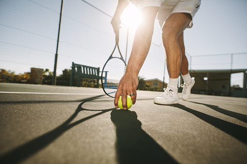Ground level view of a man picking a tennis ball on a  sunny day. Low angle view of a tennis player bending forward to pick the ball with sun in the background.