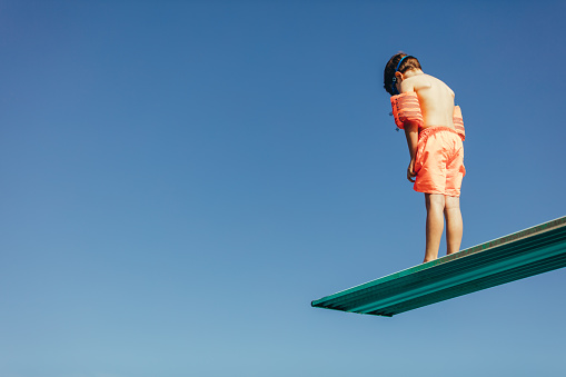 Low angle shot of boy with sleeves floats on diving board preparing for dive in the pool. Boy standing on diving spring board against sky.