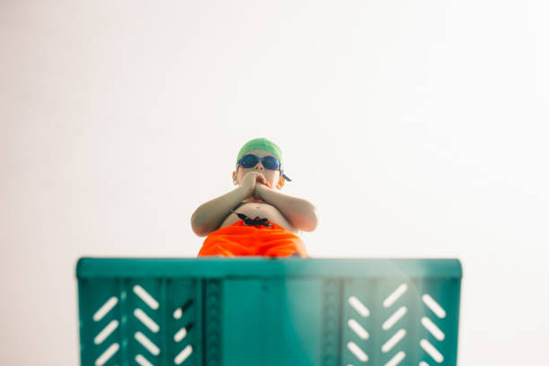 Boy on diving platform at pool Low angle view of boy in swimming trunks, swim goggles and swimming cap standing on diving board against bright sky. Boy on diving platform at pool. diving board stock pictures, royalty-free photos & images