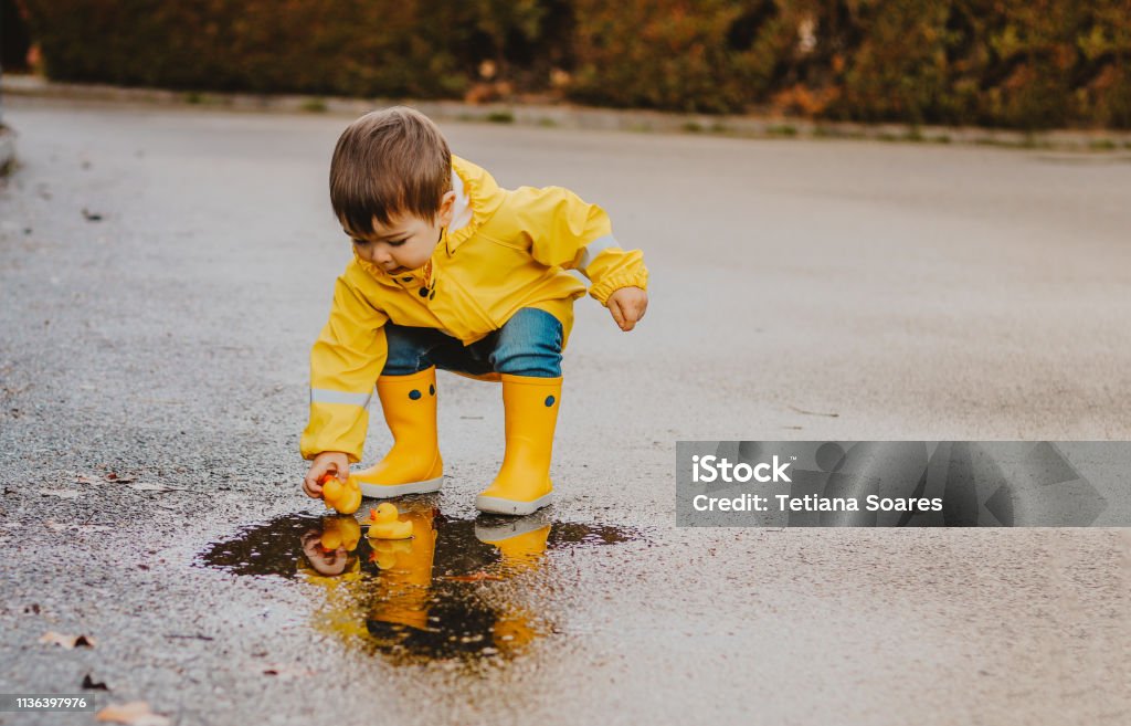 Süßer verspielter kleiner Junge in leuchtend gelbem Regenmantel und Gummistiefeln, der mit Gummienten in kleiner Pfütze am regnerischen Frühlingstag auf nasser Straßenstraße spielt. Saisonales Wetterwanderkonzept - Lizenzfrei Kind Stock-Foto