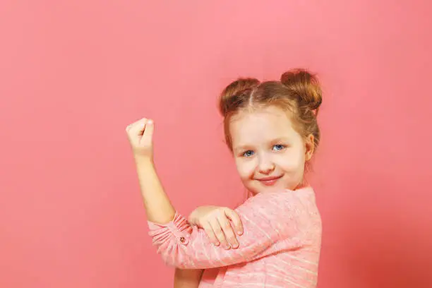 Photo of Closeup portrait of cute kid with hair buns over pink background. Child hand on biceps show how girl power