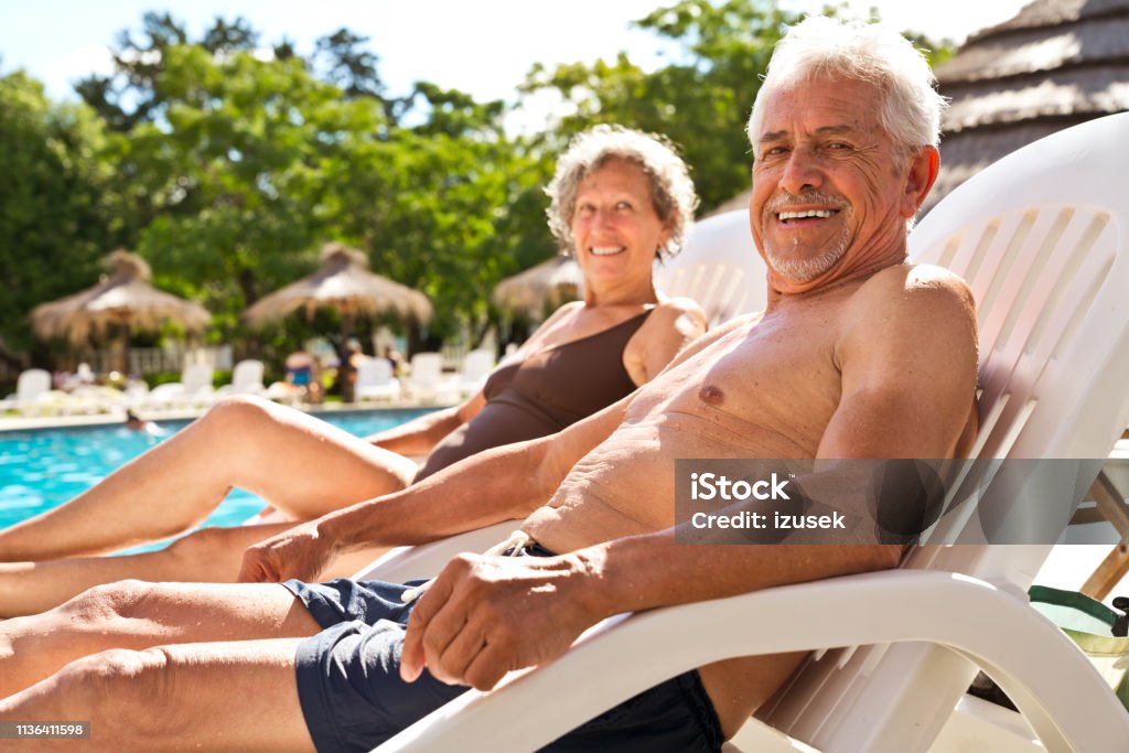 Retired couple resting on deck chairs at poolside Portrait of smiling senior man with woman resting on deck chairs. Elderly couple is relaxing at poolside on summer vacation. They are in swimwear. Swimming Pool Stock Photo