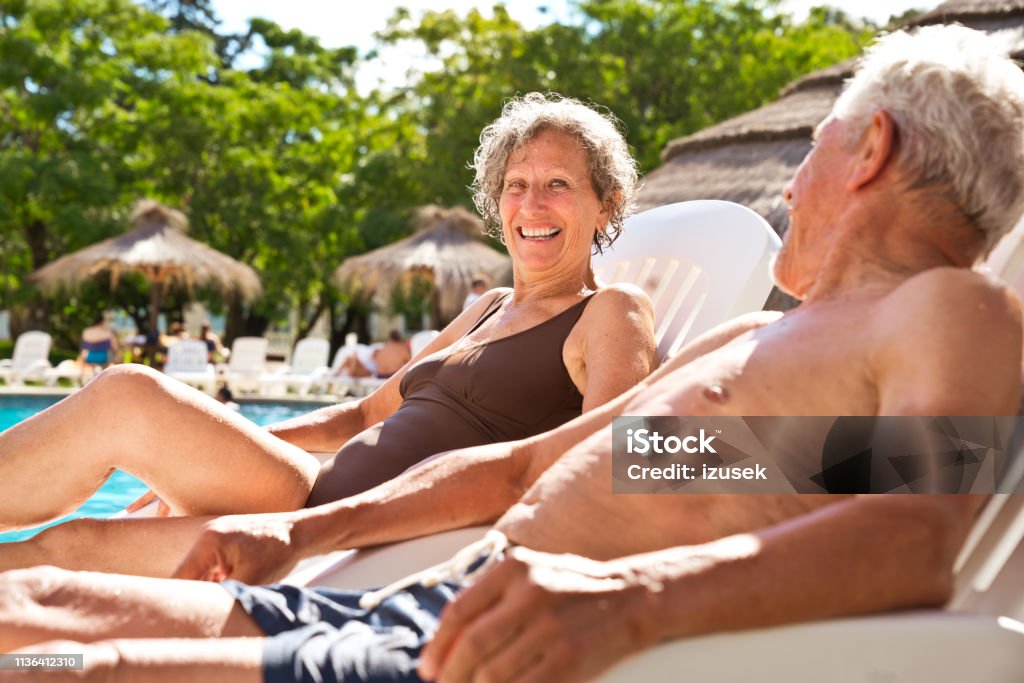 Senior couple talking while resting on deck chairs Smiling senior woman talking to man at poolside. Elderly retired couple is resting on deck chairs. They are spending leisure time on summer vacation. Poolside Stock Photo