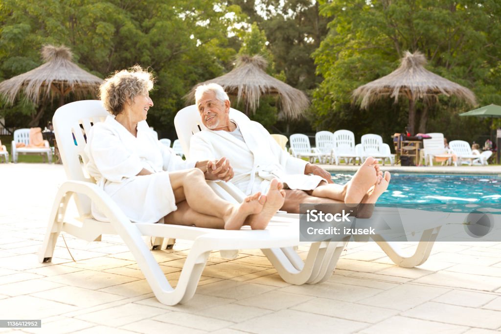 Couple holding hands while resting on deck chairs Smiling elderly couple talking while resting on deck chair. Senior man and woman are holding hands while resting by swimming pool. They are relaxing at summer vacation. 60-64 Years Stock Photo