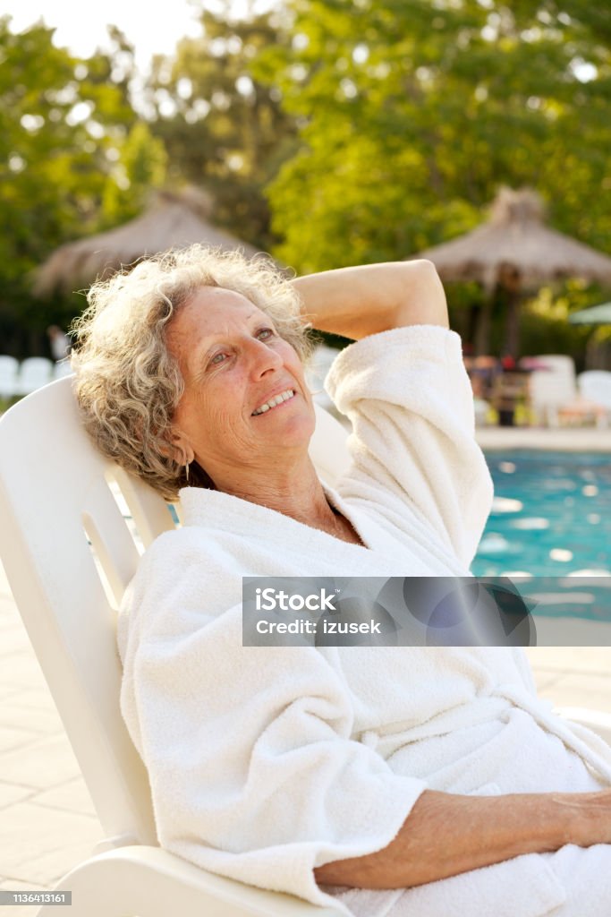 Thoughtful senior woman resting on deck chair Thoughtful smiling senior woman resting on deck chair. Elderly female is wearing bathrobe. She is looking away while relaxing at poolside. Senior Women Stock Photo