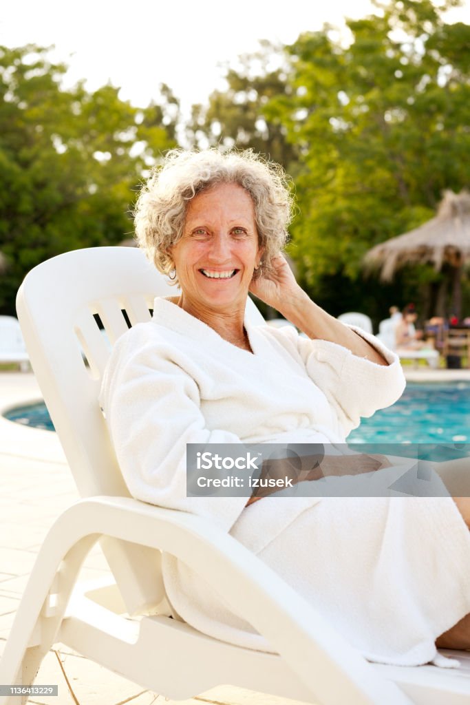 Senior woman resting on deck chair at poolside Portrait of smiling senior woman sitting on deck chair. Happy elderly female is wearing bathrobe. She is relaxing at poolside during summer vacation. 60-64 Years Stock Photo