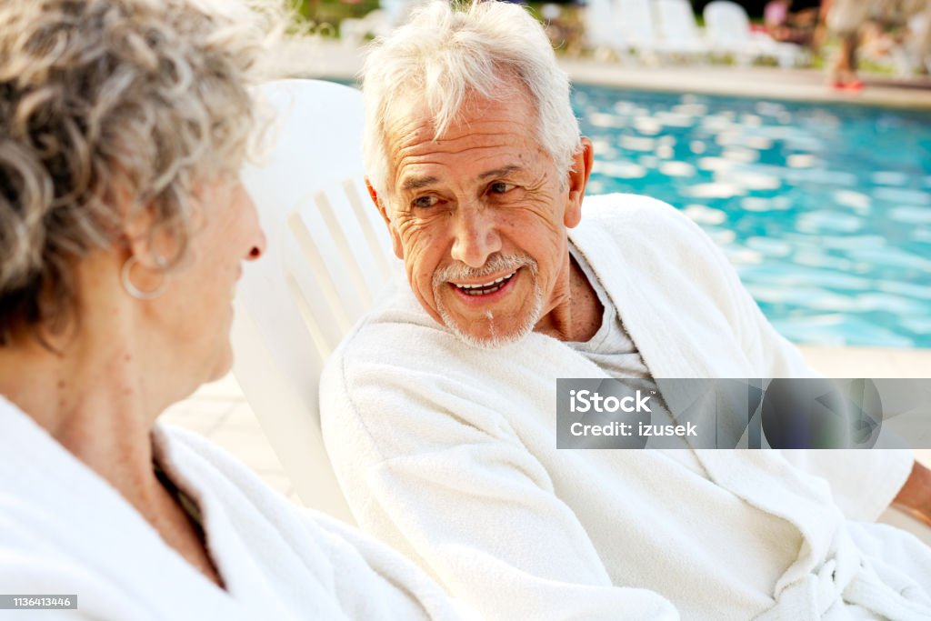 Senior couple talking while resting on deck chairs Smiling senior man talking to woman at poolside. Elderly couple is resting on deck chairs. They are spending leisure time on summer vacation. Senior Couple Stock Photo