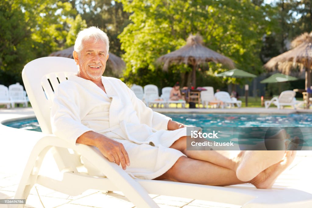 Portrait of senior man on deck chair at poolside Full length of smiling senior man sitting on deck chair. Portrait of happy elderly male is wearing bathrobe. He is relaxing at poolside during summer vacation. Deck Chair Stock Photo