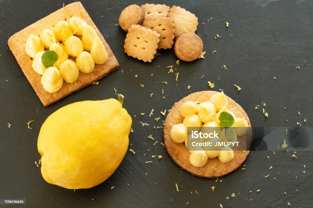 Homemade lemon tart from Menton Two partes with lemon of Menton with a lemon and small cakes placed next to it. all set on a slate with a bird's eye view. Menton Lemon has a softer taste, more intense aromas and moderate acidity. It has been protected by the European "Protected Geographical Indication" (PGI) label since 2015. Lemon - Fruit Stock Photo