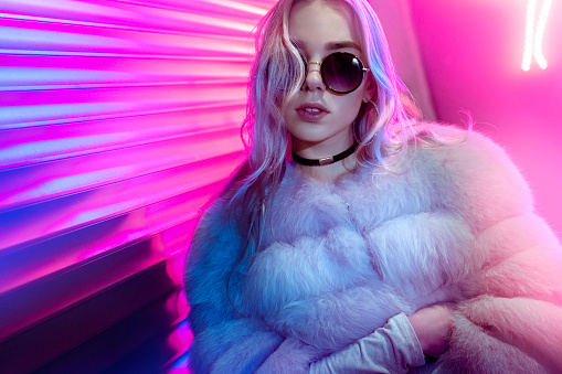 Teen hipster girl in stylish glasses and fur posing on street neon light wall background, female teenager fashion model woman with beautiful face looking at camera in city night light glow, back to 80s, portrait