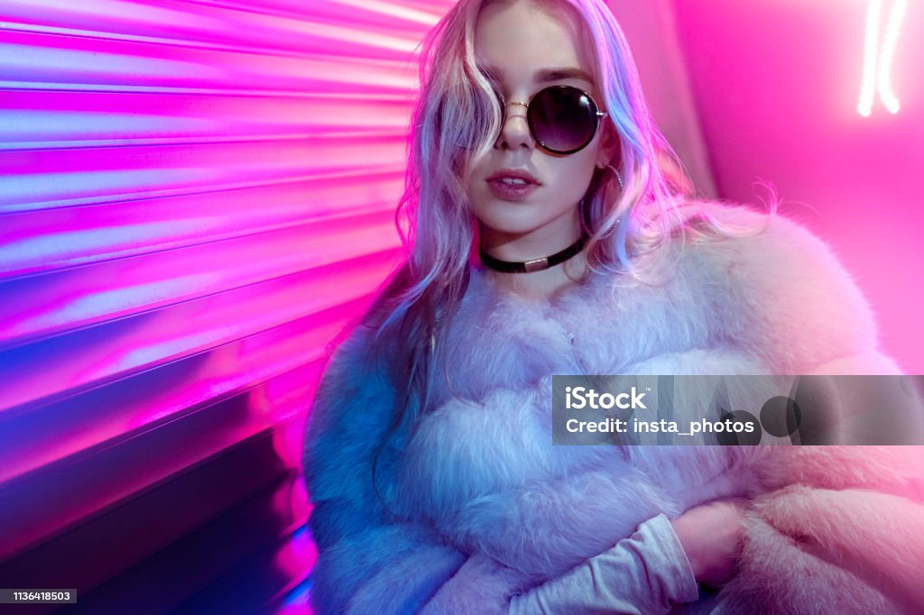 Teen hipster girl in stylish glasses and fur posing on street neon light wall background, female teenager fashion model woman with beautiful face looking at camera in city night light glow, back to 80s, portrait Teenage Girls Stock Photo