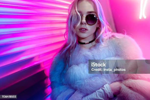 Teen Hipster Girl In Stylish Glasses And Fur Posing On Street Neon Light Wall Background Female Teenager Fashion Model Woman With Beautiful Face Looking At Camera In City Night Light Glow Back To 80s Portrait Stock Photo - Download Image Now