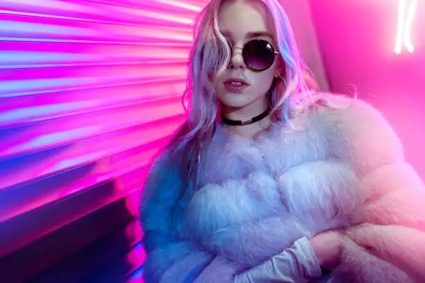 Photo of Teen hipster girl in stylish glasses and fur posing on street neon light wall background, female teenager fashion model woman with beautiful face looking at camera in city night light glow, back to 80s, portrait