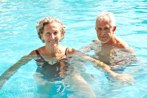 Portrait of senior couple swimming in pool. Retired elderly man and woman are enjoying in water. They are having fun in vacation.