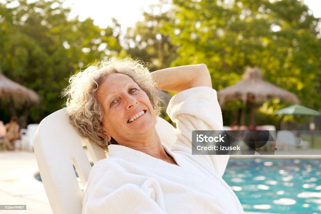 Smiling senior woman resting at poolside Portrait of smiling elderly woman resting on deck chair. Happy senior female is relaxing at poolside. She is in bathrobe during summer vacation. One Woman Only Stock Photo