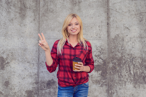 Smiling happy woman with blonde hair dressed in casual clothes is showing victory sign and holding morning coffee