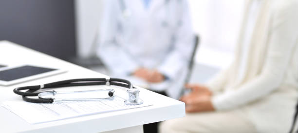 Stethoscope, clipboard with medical form lying on hospital reception desk with laptop computer and busy doctor and patient communicating at the background. Medical tools at doctor working table.Medicine concept. estetoscopio, portapapeles con forma médica acostado en la recepción del hospital con ordenador portátil y el médico ocupado y paciente de comunicación en el fondo. herramientas médicas en la mesa de trabajo del médico. concepto de medicina.
