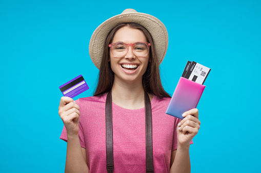 Excited young female tourist holding credit card and passport with tickets, ready to flight, isolated on blue background