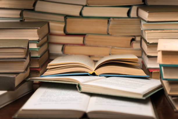 istock Education learning concept with open book and notebook on the table. Stack piles of books on reading desk and glasses in bookshelves background 1136449499