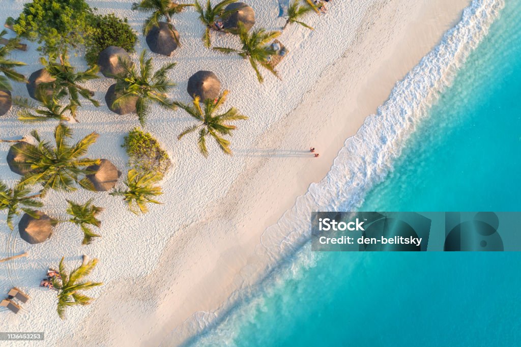 Luftaufnahme von Sonnenschirmen, Palmen am Sandstrand des Indischen Ozeans bei Sonnenuntergang. Sommerurlaub in Sansibar, Afrika. Tropische Landschaft mit Palmen, Sonnenschirmen, weißem Sand, blauem Wasser, Wellen. Top View - Lizenzfrei Strand Stock-Foto