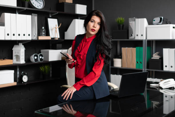 istock Elegant woman sat down on table and holding phone in office 1136476699