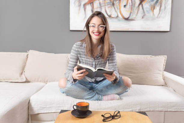 istock Young woman sitting on sofa,looking in to a camera and holding a book 1136487972