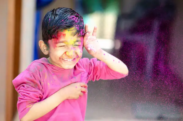 Photo of Cute Little Indian boy child with coloured face paint powder color thrown at his face during holi indian festival looking at camera