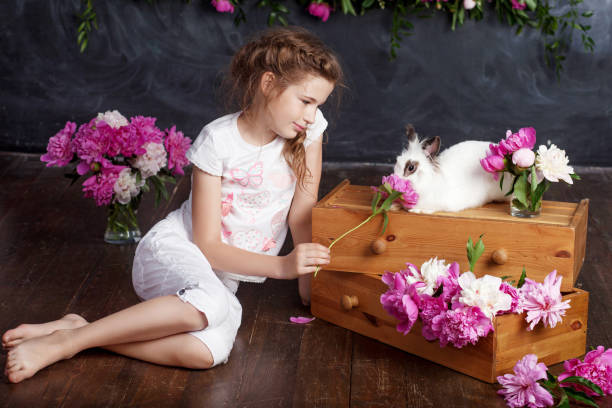 本物のウサギと遊んでいる小さな女の子。花の背景にイースターの子供と白のバニー。子供とペットが遊びます。動物や子供のための楽しさと友情。 - easter easter bunny fun humor ストックフォトと画像
