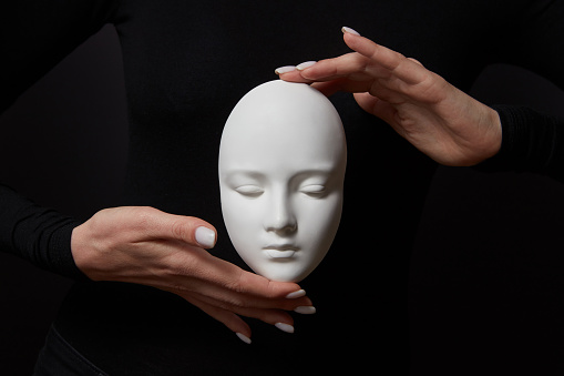 White plaster mask face is holding woman's fingers on a black background, copy space. Concept social psychological masks