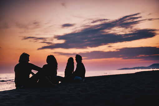 Silhouettes of four friends relaxing on the beach at dusk. Copy space.