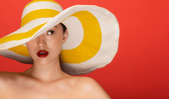 Close up portrait of attractive young asian woman wearing summer hat staring at side against red background.