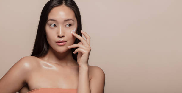 Woman applying beauty product on face Young Korean woman applying moisturizer to her face over beige background. Young woman with cosmetic cream on a cheek and looking away. asian american model stock pictures, royalty-free photos & images