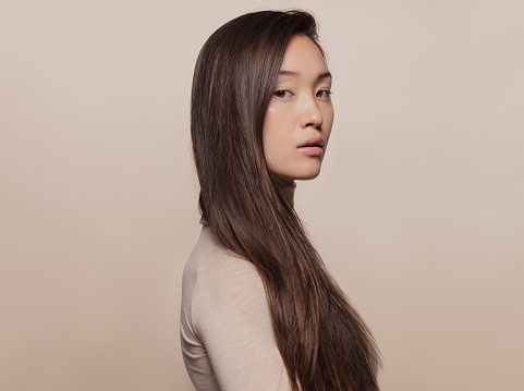 Portrait of beautiful young woman with long brown hair standing against beige background. Asian woman with a long straight hair looking at camera.