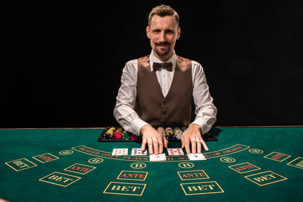 il ritratto di un croupier tiene le carte da gioco, le fiches da gioco sul tavolo. sfondo nero - croupier foto e immagini stock