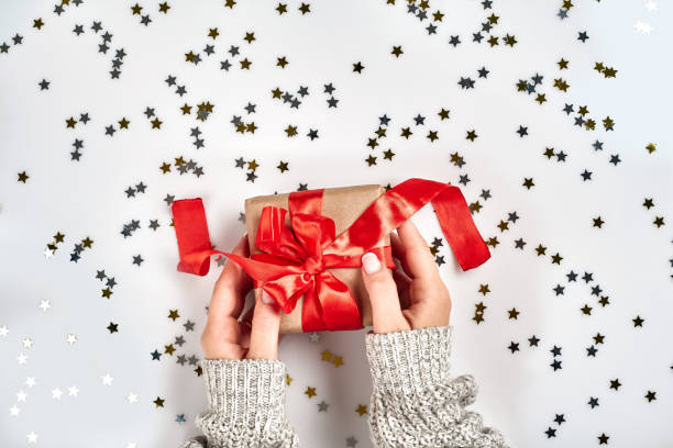 istock Female hands holding present with red bow on white background with delicate wavy ribbons and metallic star shaped confetti. Flat lay style 1136570346