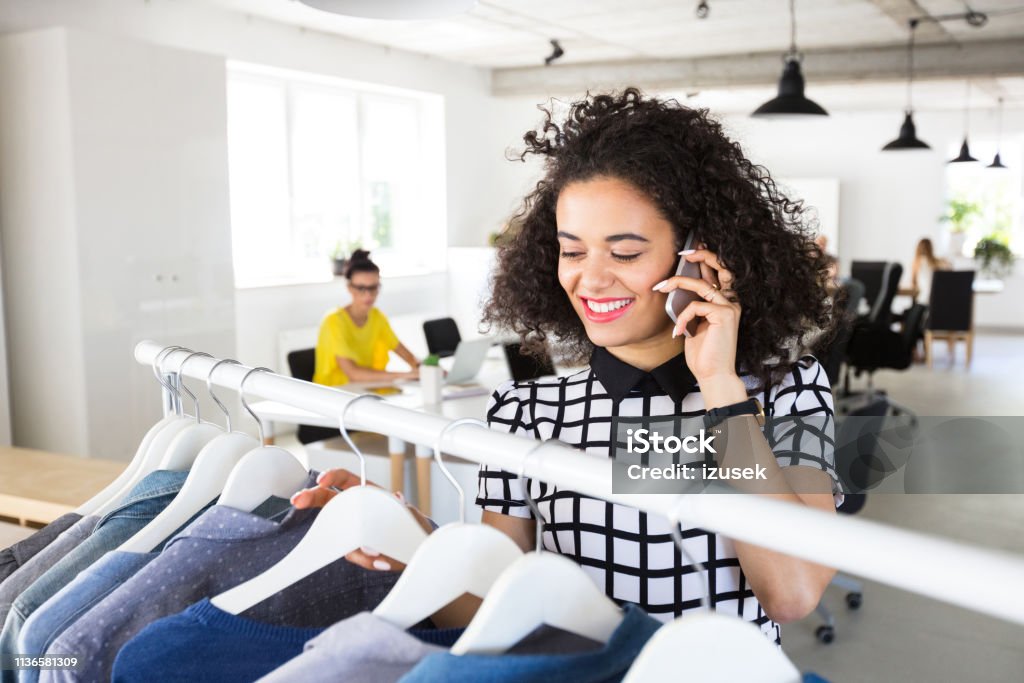Fashion designer working in modern workplace Smiling young woman looking at the clothes on the rack and talking on mobile phone in office Busy Stock Photo