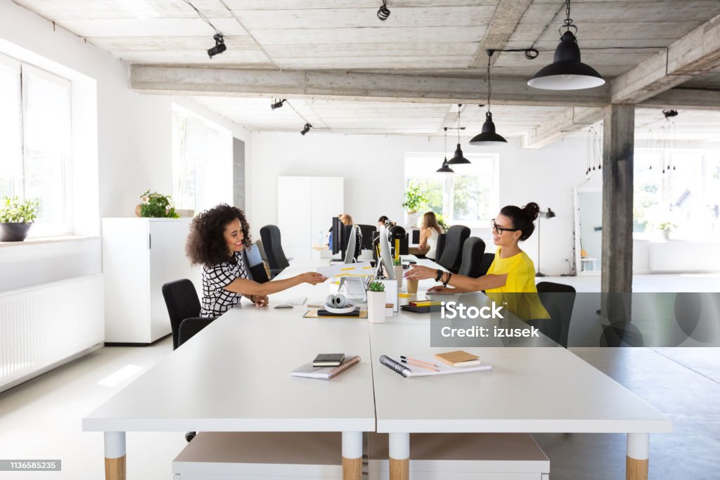 Businesswomen working at modern workplace Shot of young businesswomen working on computers at modern workplace, passing a document. Office Stock Photo