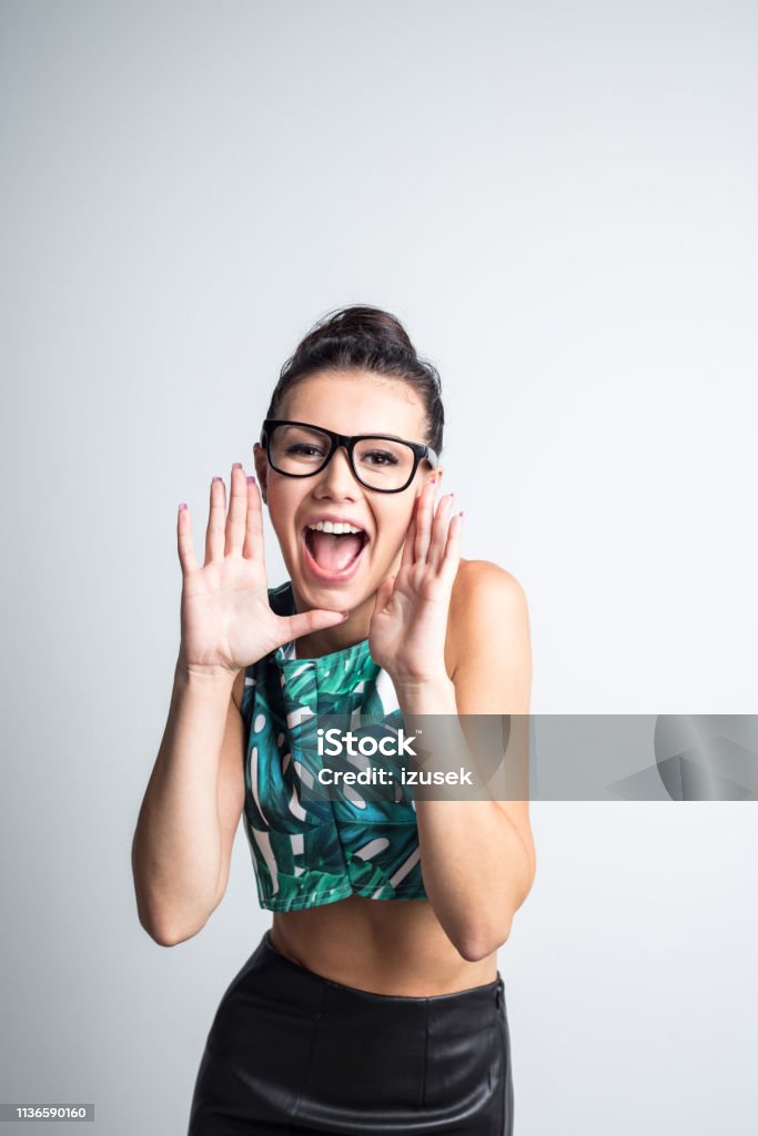 Excited young woman shouting Portrait of excited young woman shouting with hands around mouth against grey background Announcement Message Stock Photo
