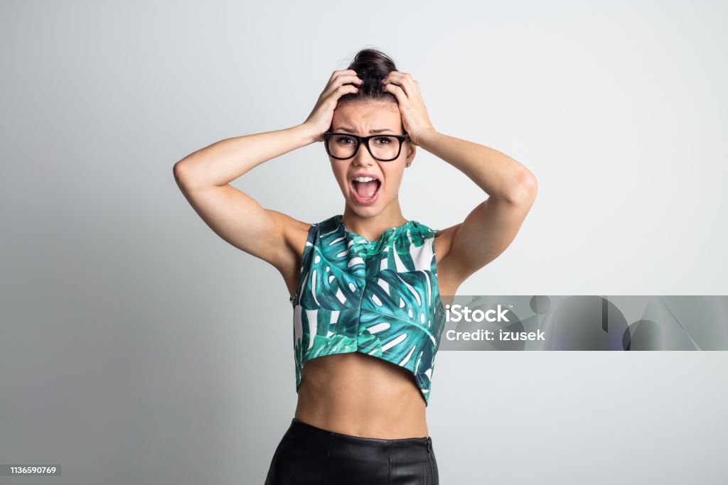 Beautiful girl screaming with hands on head Portrait of young woman in stylish outfit looking shocked, screaming with hand in hand against grey background 20-24 Years Stock Photo