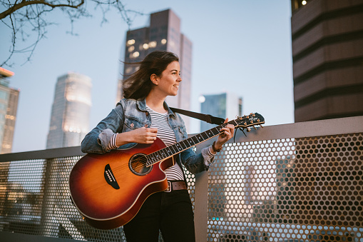 A young Caucasian woman has fun playing her acoustic guitar outside in downtown Los Angeles, California.  Shot at dusk, the city lights illuminating the scene behind her.