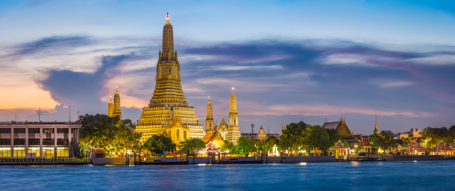 The iconic spire of Wat Arun spotlit against the blue dusk sky across the Chao Phraya River in the heart of Bangkok, Thailand’s vibrant capital city.