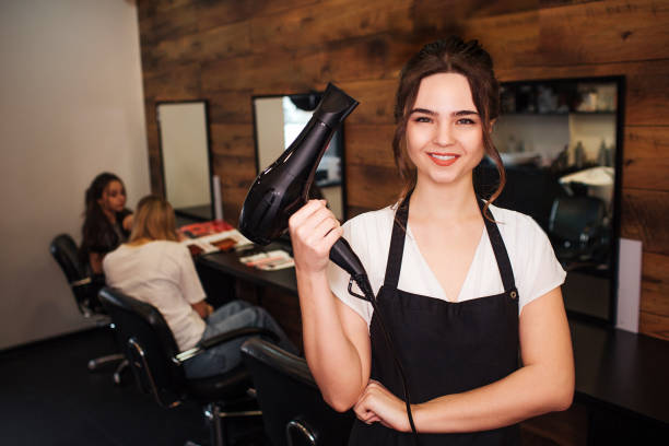 portret de peluquero sonriente en salón de belleza. hermosa mujer en delantal negro mirando a la cámara y sosteniendo secador de pelo profesional. concepto de belleza y personas - barbero peluquero fotografías e imágenes de stock