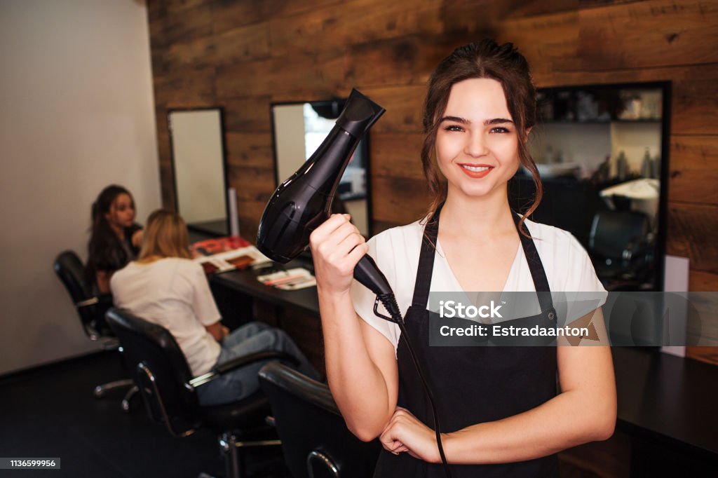 Portret of smiling hairdresser in beauty salon. Beautiful woman in black apron looking at camera and holding professional hair dryer. Beauty and people concept Portret of smiling hairdresser in beauty salon. Beautiful woman in black apron looking at camera and holding professional black hair dryer. Beauty and people concept Hairdresser Stock Photo