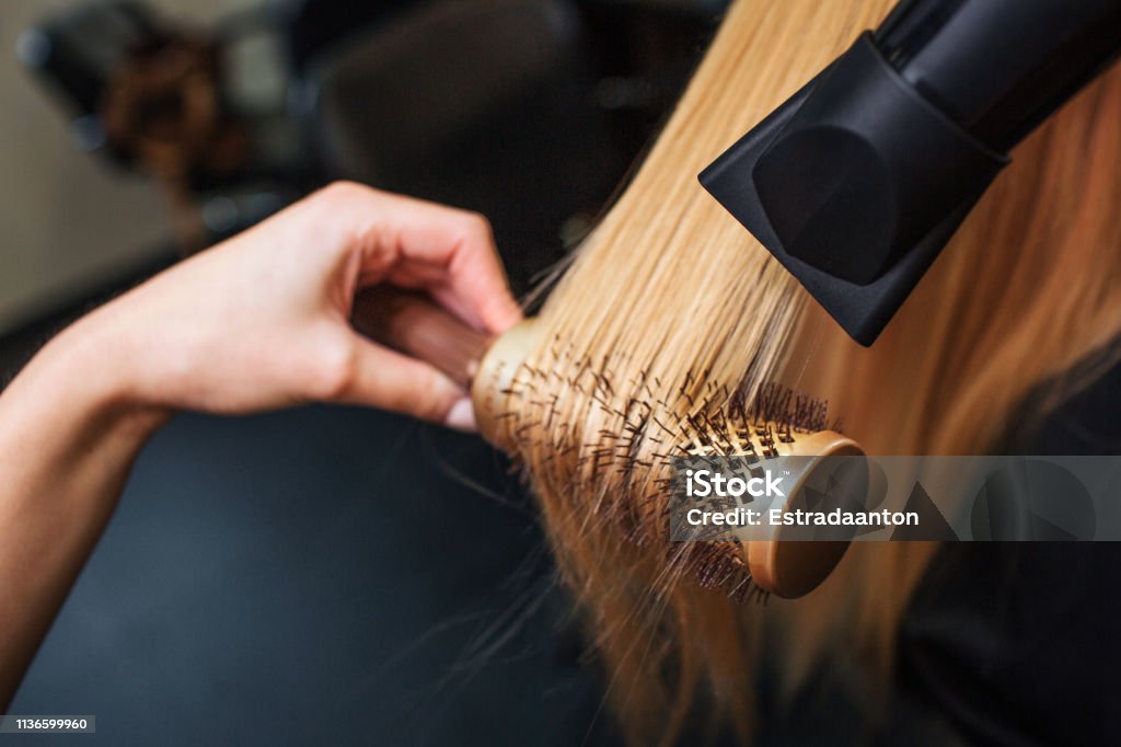 Female client in beauty salon. Close-up of hairdressers hand drying blond hair with hair dryer and round brush, doing new hairstyle Female client in beauty salon. Close-up of hairdressers hand drying blond hair with hair dryer and round brush, doing professional hairstyle Hair Salon Stock Photo